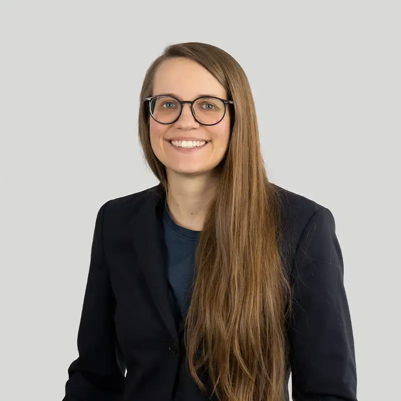 Eine Frau in einem schwarzen Blazer mit blauem Shirt, Nerdbrille und langen braun-blonden Haaren. Sie lächelt in die Kamera. 